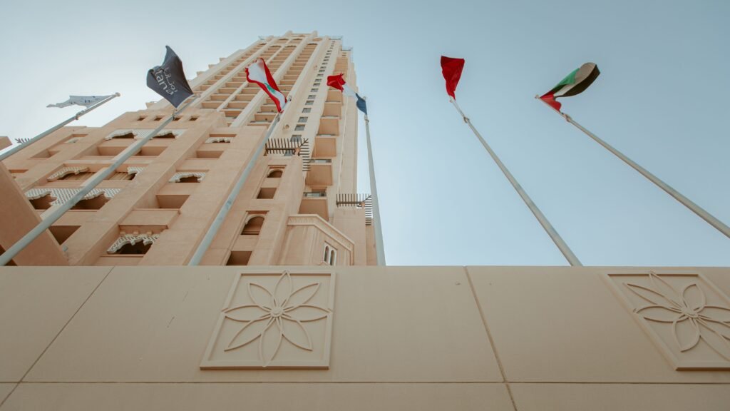 A low-angle view of a tall, beige high-rise building with Middle Eastern architectural details and international flags flying against a clear sky.